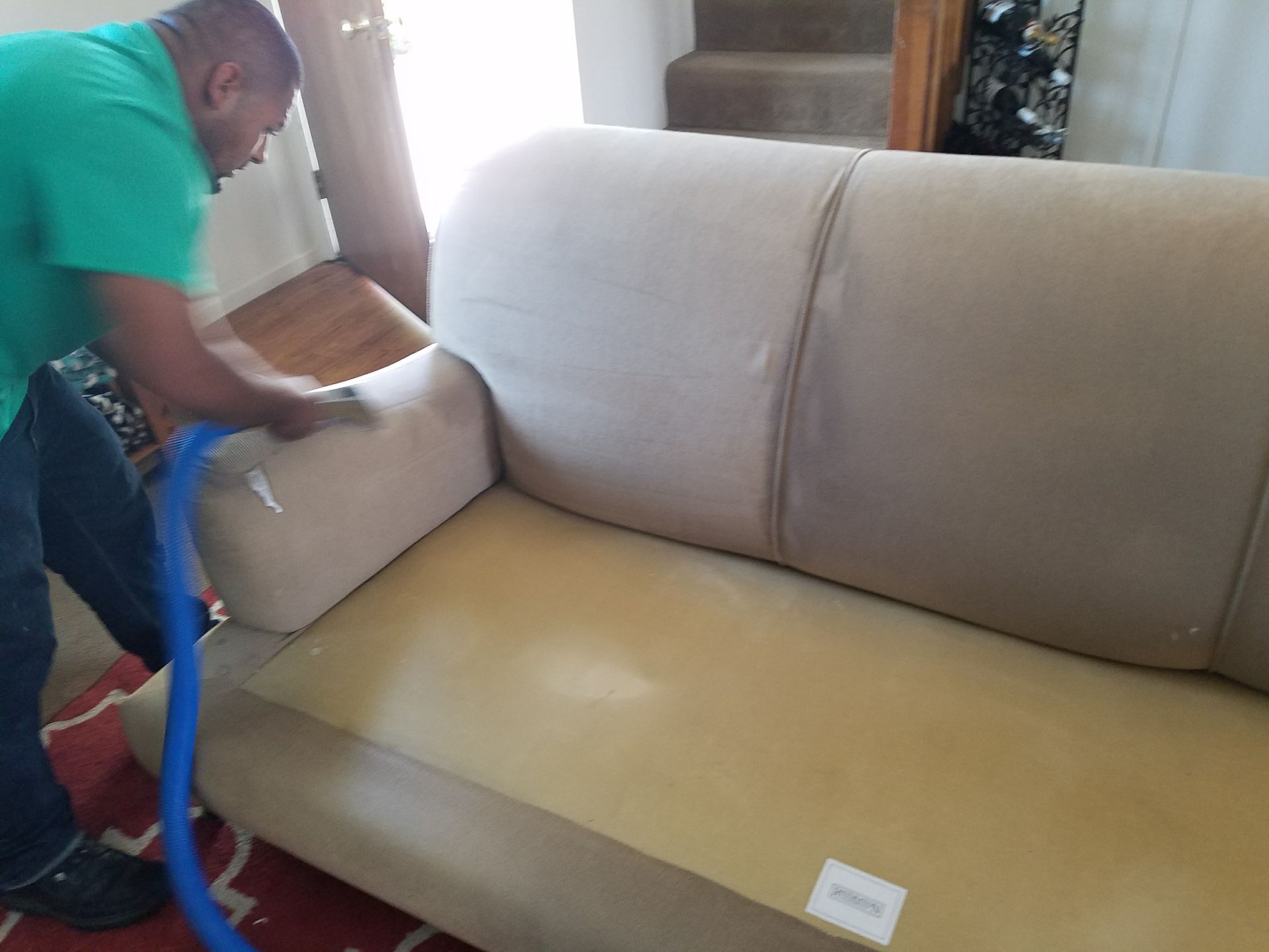 Person cleaning a beige sofa with a blue hose attachment.