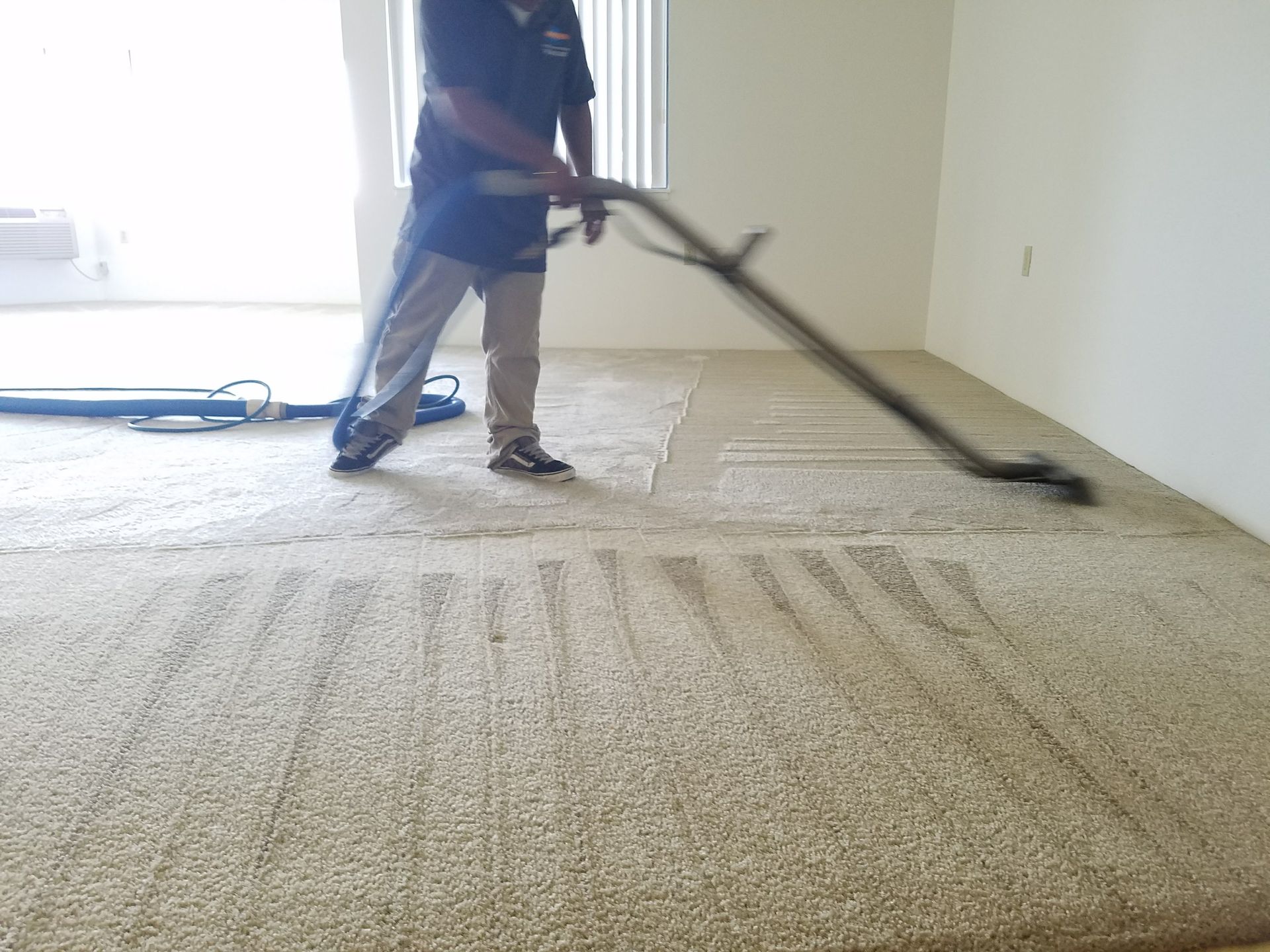 Person vacuuming a beige carpet in an empty room with a wall-mounted air conditioner.