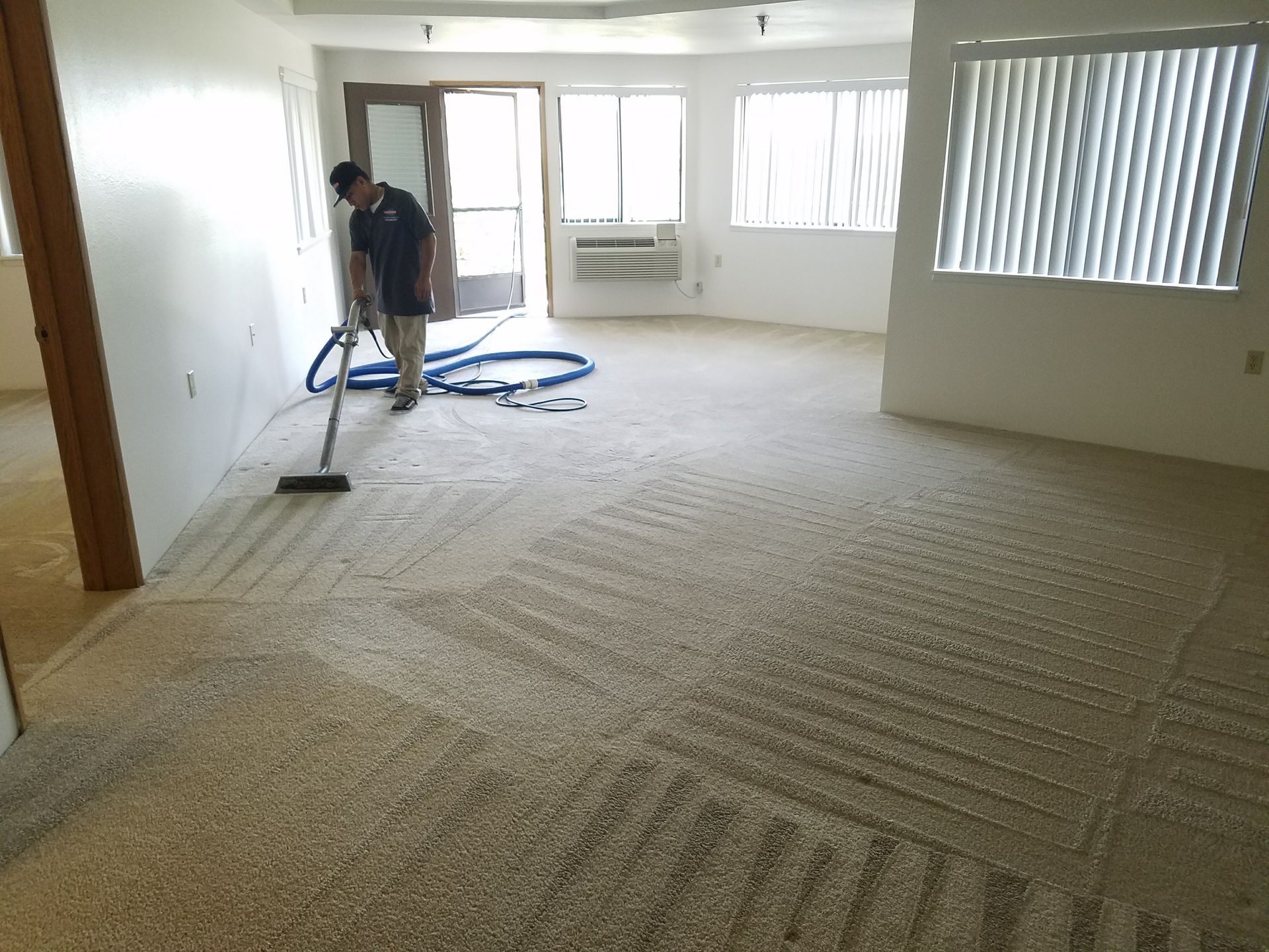 Worker vacuuming carpet in a bright empty room with large windows and doors